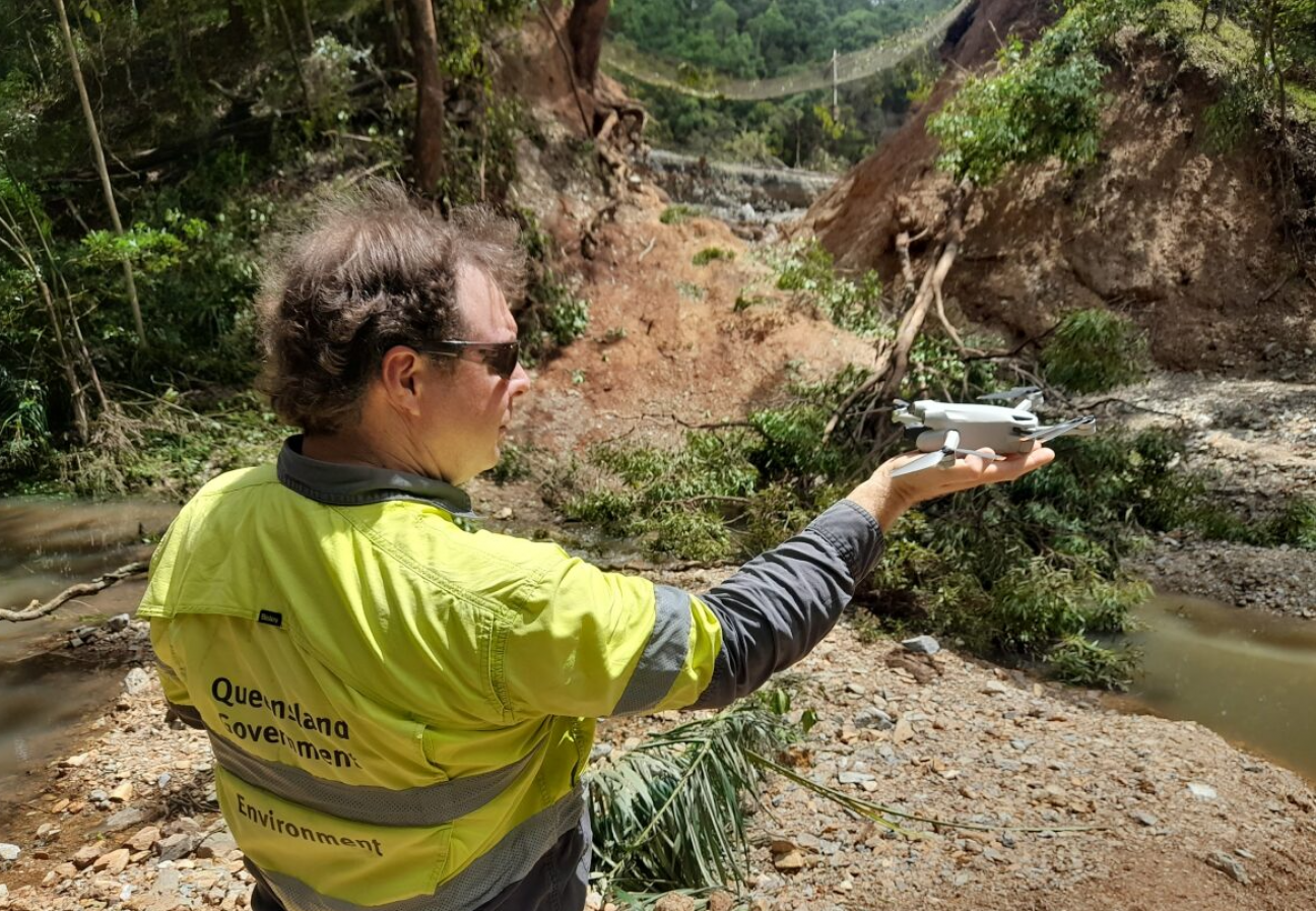 Photo of a person holding a drone while standing in a flood affected bushland.